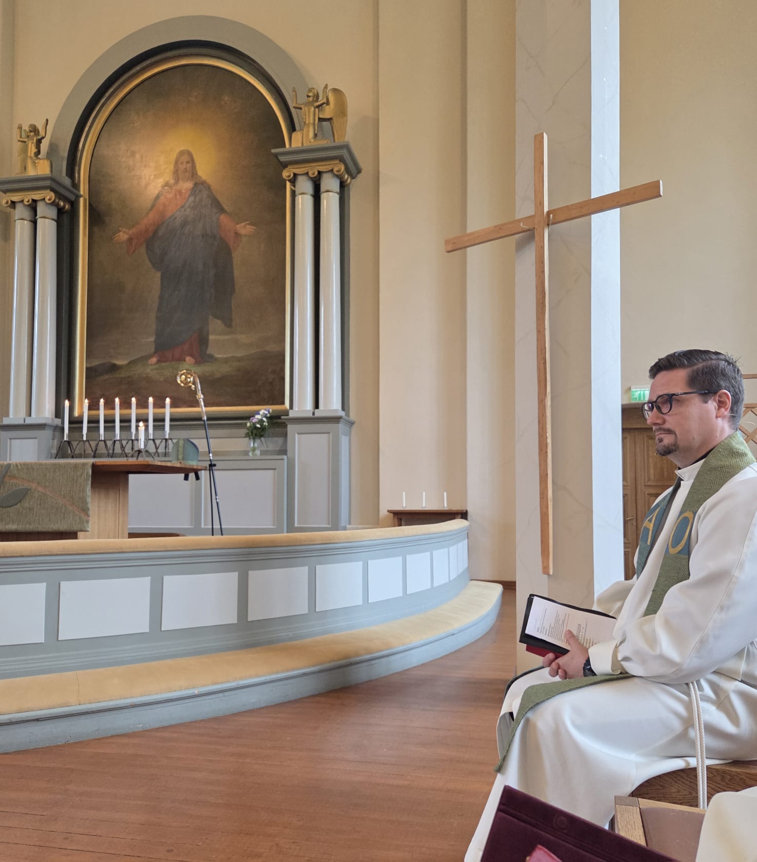 Kirkkoherra Tuomas Niskanen istuu virsikirja kädessä. Taustalla alttaritaulu ja risti.