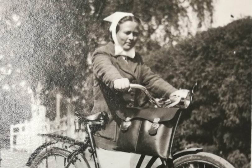 Kuva 1930- tai 1940-luvulta: Nainen, jolla polkupyörä, jonka sarvessa roikkuu salkku. Naisella on hilkka pä...