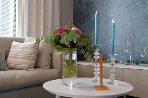 Olohuoneen pöydällä on kaksi kynttilää, kukkia maljakossa ja pieni risti.